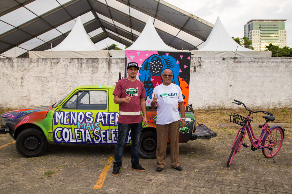 Dois homens fazendo o gesto de polegar levantado em frente a um carro com as palavras "Menos aterros mais coleta seletiva"
