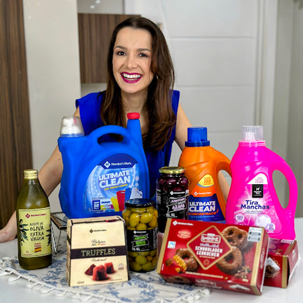 Uma mulher sorridente atrás de uma mesa com diversos bolos realistas em formato de itens de supermercado.
