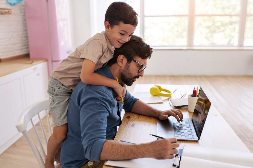 Um homem trabalhando no laptop enquanto o filho o abraça.